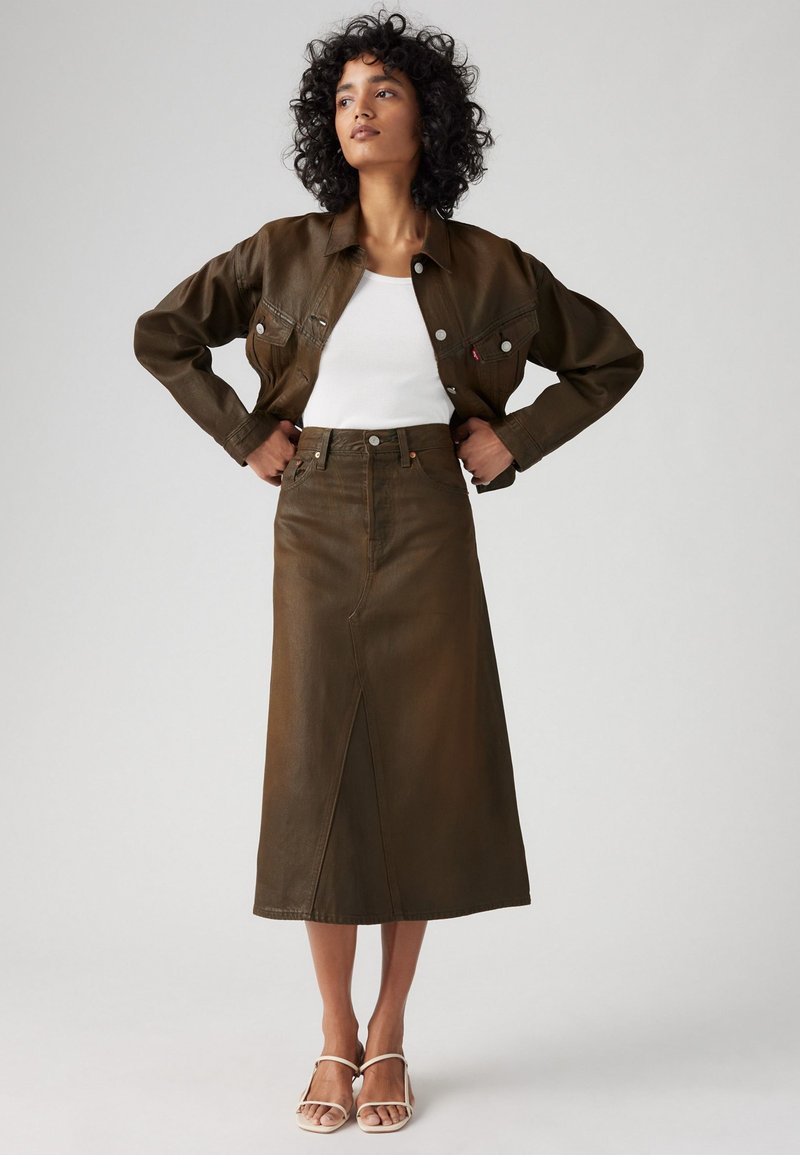 Chaqueta de cuero marrón y falda larga a juego con una abertura frontal. Camiseta blanca por debajo. Sandalias beige con tiras completan el conjunto.