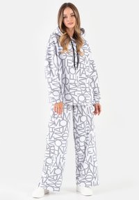 Kobieta ubrana w białą bluzę z kapturem i szerokie spodnie ozdobione czarnymi abstrakcyjnymi literami, stojąca na białym tle.