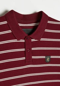 Bordowa koszulka polo z białymi poziomymi paskami, z prążkowanym kołnierzykiem i dwoma guzikami. Posiada czarne logo w kształcie tarczy z emblematem lwa.