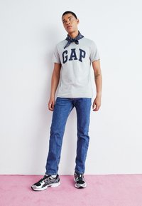 Camiseta de algodón gris con el logo "GAP" en azul marino, combinada con jeans de mezclilla azul. Zapatillas negras con acentos plateados. El modelo posa frente a un fondo rosa.