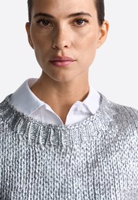 Srebrny sweter z dzianiny o fakturowanym wykończeniu, szerokim dekolcie i prążkowanych detalach, nałożony na świeżą białą koszulę. Gładki materiał.