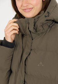 Olivgrüne Steppjacke mit hohem Kragen, Druckknöpfen und einem mattierten Logo. Sie hat eine weiche Oberfläche und einen entspannten Schnitt.