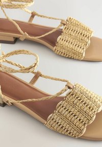 Vlakke sandalen met een geweven rieten bovenkant in beige, romige banden en een kleine blokhak. Kenmerkt zich door een kruislings vetersysteem met een textuurdesign.