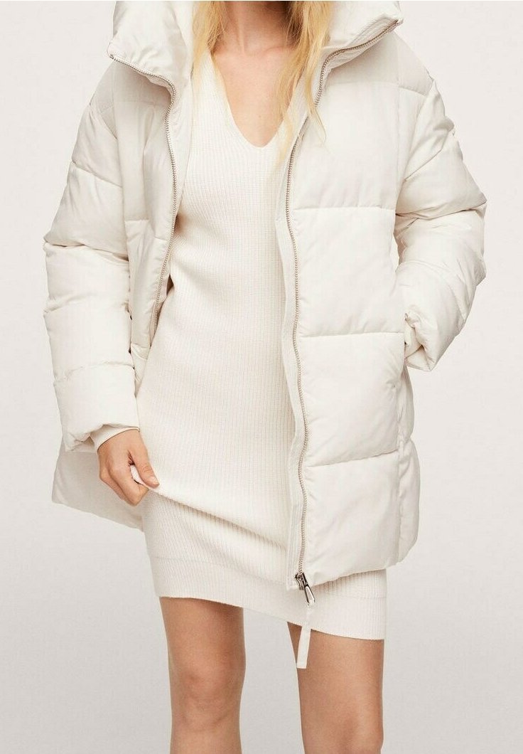 Femme portant une robe en maille côtelée beige clair et une doudoune crème avec un col montant et une fermeture éclair à l'avant.