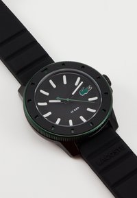 Montre Lacoste noire avec un boîtier rond, une lunette verte et des index blancs. Le bracelet en caoutchouc présente une texture striée et un logo en relief. Résistante à l'eau jusqu'à 10 bars.