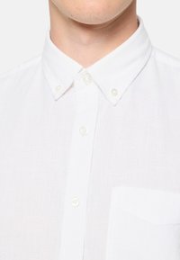 Camisa blanca de botones confeccionada en lino ligero, con un cuello clásico, cierre delantero de botones y un bolsillo en el pecho. Textura suave.