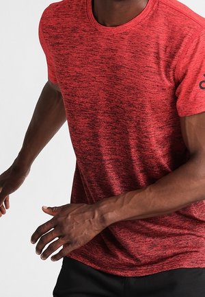 Mężczyzna w czerwonej, teksturowanej koszulce z krótkim rękawem i czarnych spodniach sportowych, pozujący z lekko zgiętymi ramionami na białym tle.