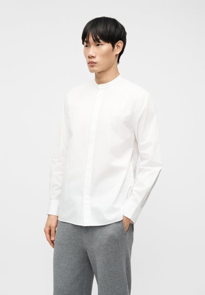 Νεαρός άνδρας με μαύρα μαλλιά, φορεί λευκό πουκάμισο με μανικετόκουμπα μανταρίνι και γκρι παντελόνι, στέκεται με το ένα χέρι στην τσέπη μπροστά σε απλό φόντο.