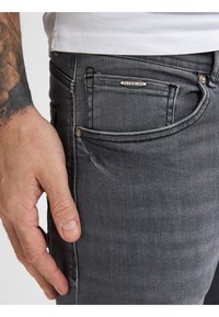 Jeans de mezclilla de gris oscuro con un corte ajustado, que presentan un bolsillo frontal con una etiqueta de la marca y detalles de costura sutiles. Mano descansando en el costado.