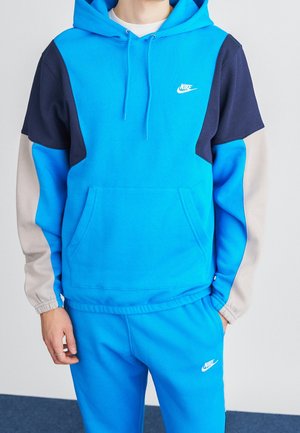 Person iført en knaldblå Nike-hættetrøje med ærmer i marineblåt og beige farveblokke samt matchende blå Nike-sweatpants mod en ensfarvet baggrund.