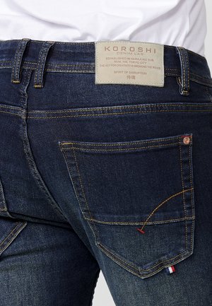 Vaqueros de denim azul oscuro con costuras amarillas, una etiqueta KOROSHI y detalles de acento que incluyen una pequeña bandera roja, blanca y azul cerca del bolsillo.