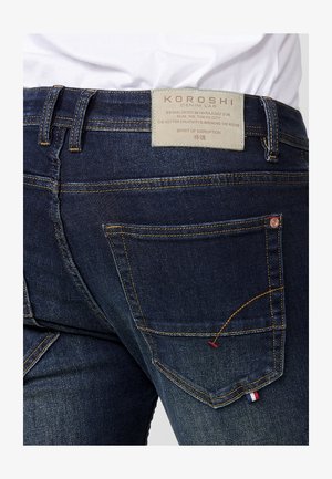 Tumedast sinised denim püksid kollaste õmblustega, KOROSHI silt ja aktsentdetailid, sealhulgas väike punane, valge ja sinine lipp tasku lähedal.
