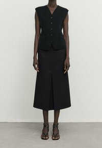 Gilet noir sans manches avec un décolleté en V profond et des boutons sur le devant, associé à une longue jupe noire avec une fente à l'avant et un design minimaliste.