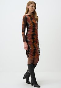 Figurerekkende jurk in oranje, bruin en zwart, met lange mouwen en een geplooide textuur, gecombineerd met kniehoge zwarte laarzen.