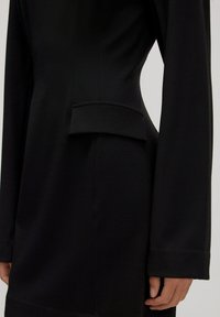 Close-up van een zwart op maat gemaakte blazer met lange mouwen, met een gestructureerde taille en een horizontaal flapdetail op de zijzak.