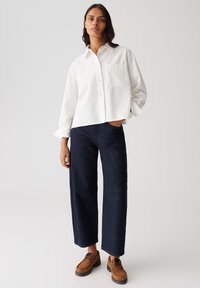 Vrouw in een loszittende witte blouse met knopen, donkere wijde jeans en bruine veterschoenen, staand met één hand in de zak.