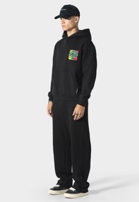 Sudadera negra con un patrón colorido de hojas en el bolsillo del pecho, combinada con pantalones de chándal negros a juego y zapatillas negras con suelas blancas.