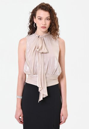 Donna con capelli ricci che indossa una camicetta beige lucida senza maniche con un fiocco al collo e una gonna nera a vita alta.