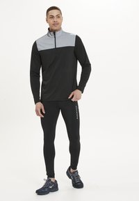 Sort atletisk langærmet top med et gråt brystpanel, kvart-zip krave; sammen med sorte tætsiddende bukser og mørke sneakers.