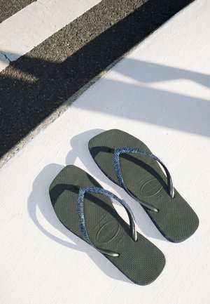 Paar dunkelgrüne Flip-Flops mit blauen glitzernden Riemen, die auf weißem Pflaster neben einer Asphaltstraße mit Schatten in der Nähe liegen.
