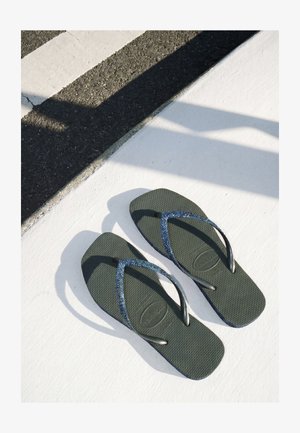 Paar dunkelgrüne Flip-Flops mit blauen glitzernden Riemen, die auf weißem Pflaster neben einer Asphaltstraße mit Schatten in der Nähe liegen.