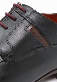 Chaussure en cuir noir avec des lacets bordeaux, une surface texturée, des détails cousus et un patch logo marron sur le côté. Design discret.