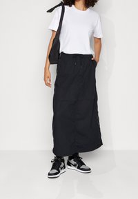 Svart maxikjol med dragsko i midjan, kombinerad med en vit t-shirt med kort ärm och svarta sneakers. Bär en liten svart axelväska.