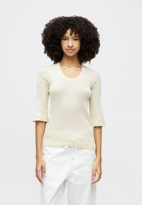 Haut en maille beige clair avec un décolleté arrondi et des manches courtes évasées. Porté avec un pantalon blanc, présentant une coupe décontractée et une taille à cordon de serrage.