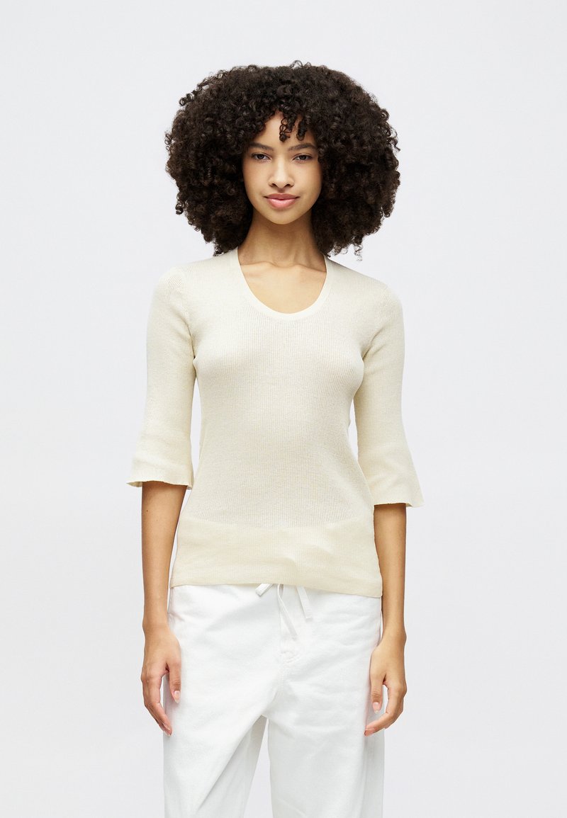 Haut en maille beige clair avec un décolleté arrondi et des manches courtes évasées. Porté avec un pantalon blanc, présentant une coupe décontractée et une taille à cordon de serrage.