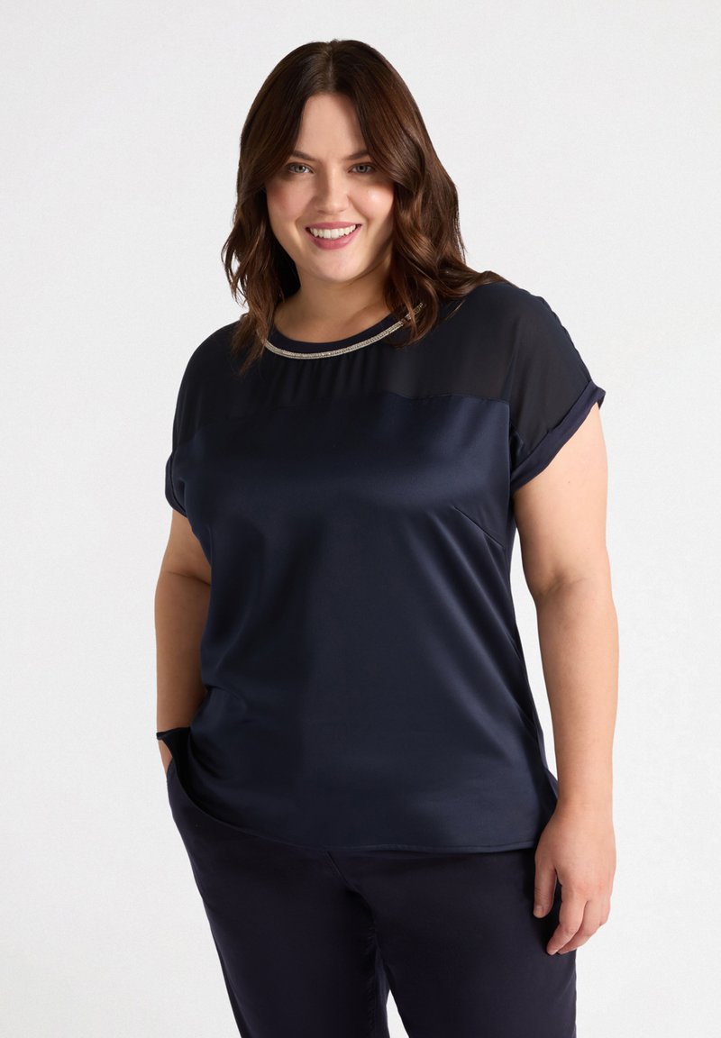 Bluză bleumarin cu o textură netedă, mâneci scurte și un panou transparent la umeri. Are un finisaj metalic în jurul gulerului.