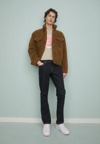 Bruine fleecejas met twee borstzakken, beige graphic t-shirt, donkere jeans en witte sneakers met roze accenten. Minimalistische achtergrond.