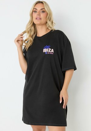 Kvinde med langt blond hår bærer en løs sort kjole med "IBIZA LA ISLA" og en blå palmeillustration på brystet mod en ensfarvet hvid baggrund.