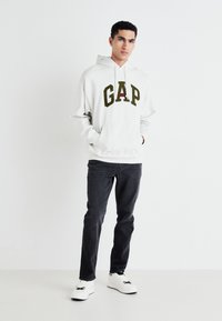Hanorac alb cu logo verde "GAP", buzunar frontal, pe un fundal simplu, asortat cu blugi și adidași albi, ținută casual.