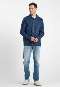 Navyblauwe katoenen jack met knopenfront, puntkraag en borstzak, gecombineerd met lichte denim jeans en witte sneakers.