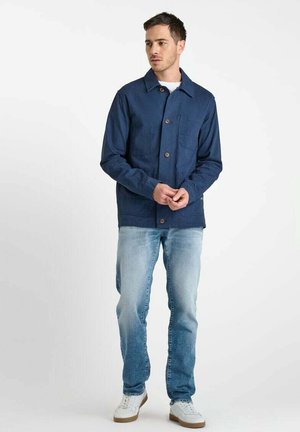 Navyblauwe katoenen jack met knopenfront, puntkraag en borstzak, gecombineerd met lichte denim jeans en witte sneakers.