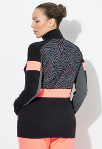 Donna con i capelli raccolti in un bun, che indossa una giacca nera e grigia con un motivo a zebra neon rosa e pantaloni abbinati in neon rosa, che si trova di profilo contro un muro bianco.