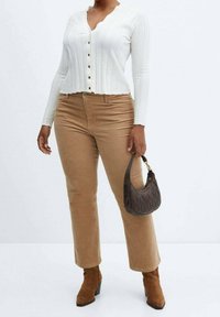 Femme portant un cardigan blanc côtelé à boutons, pantalon beige, bottines en daim marron, tenant un petit sac à main noir matelassé sur le côté.