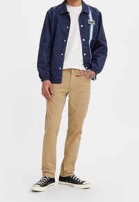 Coupe-vent bleu marine avec une bande bleue et le logo Levi's, t-shirt blanc, pantalon kaki et baskets noires avec semelles blanches.