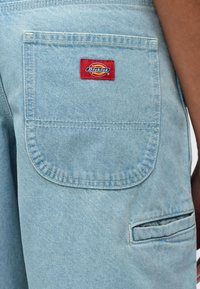 Ανοιχτό μπλε τζιν σορτς με πίσω τσέπη που έχει λογότυπο "Dickies" σε κόκκινο και κίτρινο. Ορατές ραφές και πλαϊνή τσέπη εργαλείων.