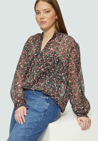 Durchscheinende schwarze Bluse mit einem Blumenmuster in Pink und Weiß, mit V-Ausschnitt und langen Puffärmeln, kombiniert mit blauen Jeans.