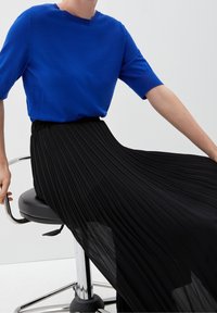 En person sitter i en svart snurrstol, klädd i en lysande blå kortärmad topp och en svepande svart plisserad kjol.