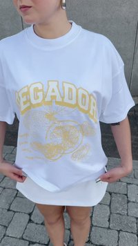 White short-sleeve T-shirt featuring bold yellow text "LEGADOR" and fruit illustrations. Soft fabric with a relaxed fit.