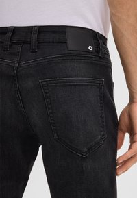 Jeans in denim nero con una texture liscia, dotati di una tasca posteriore, un'etichetta in pelle in vita e un motivo leggermente sbiadito.