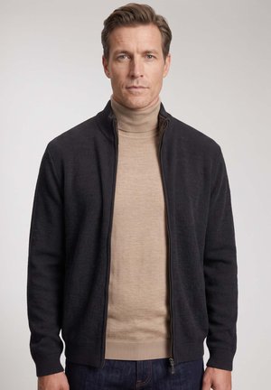 Mann mit kurzen braunen Haaren, trägt einen beigen Rollkragenpullover und einen schwarzen Pullover mit Reißverschluss, blickt mit neutralem Gesichtsausdruck nach vorne vor einfachem Hintergrund.