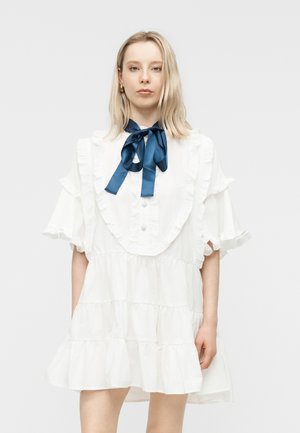 Vaaleatukkainen nainen pukeutuneena valkoiseen röyhelöhihalliseen mekkoon, jossa on suuret laivastonsiniset silkkirusetit kaulassa, seisoo yksivärisen taustan edessä.