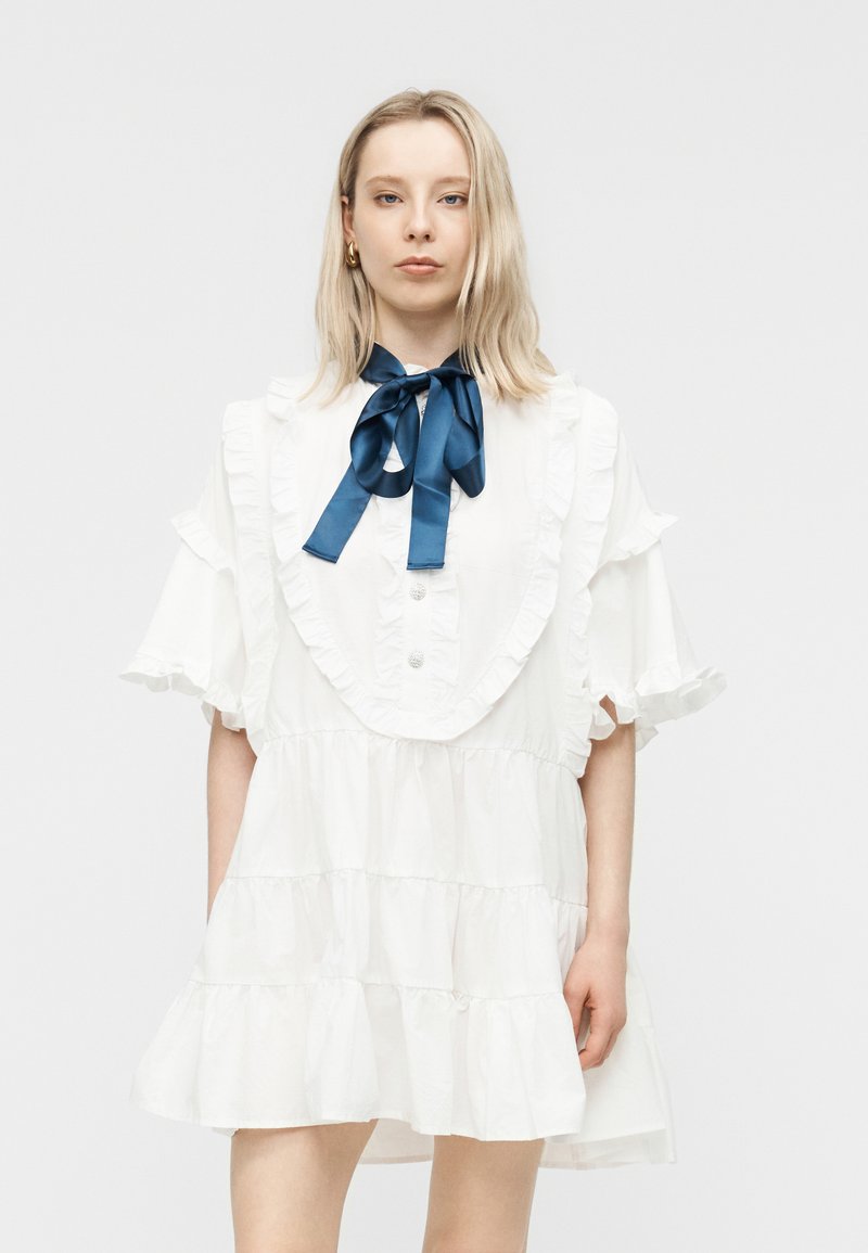 Γυναίκα με ξανθά μαλλιά φοράει λευκό φόρεμα με βολάν και φουσκωτά μανίκια, με μια μεγάλη μεταξωτή μπλε σκούρα φιόγκο δεμένη στον λαιμό, στέκεται μπροστά σε απλό φόντο.