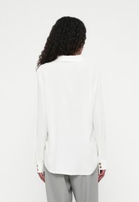 Langärmlige weiße Bluse aus glattem Stoff, mit klassischem Kragen, abgerundetem Saum und Knopfdetails an den Manschetten.