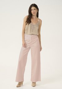 Béžová tielko s čipkovým výstrihom, v kombinácii s svetloružovými širokými nohavicami. Modelka má na sebe béžové členkové topánky na podpätku. Jednoduchý, elegantný outfit.
