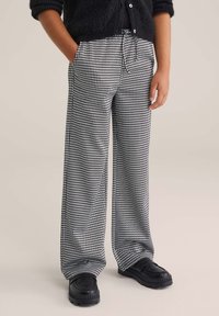Hlače s črno-belimi houndstooth vzorci, ohlapnega kroja, z vrvico na pasu, stranskimi žepi in v kombinaciji s črnimi čevlji.