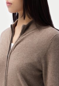 Ljusbrun zip-up tröja i mjukt tyg, med hög krage, ribbade detaljer och en elegant silhuett.